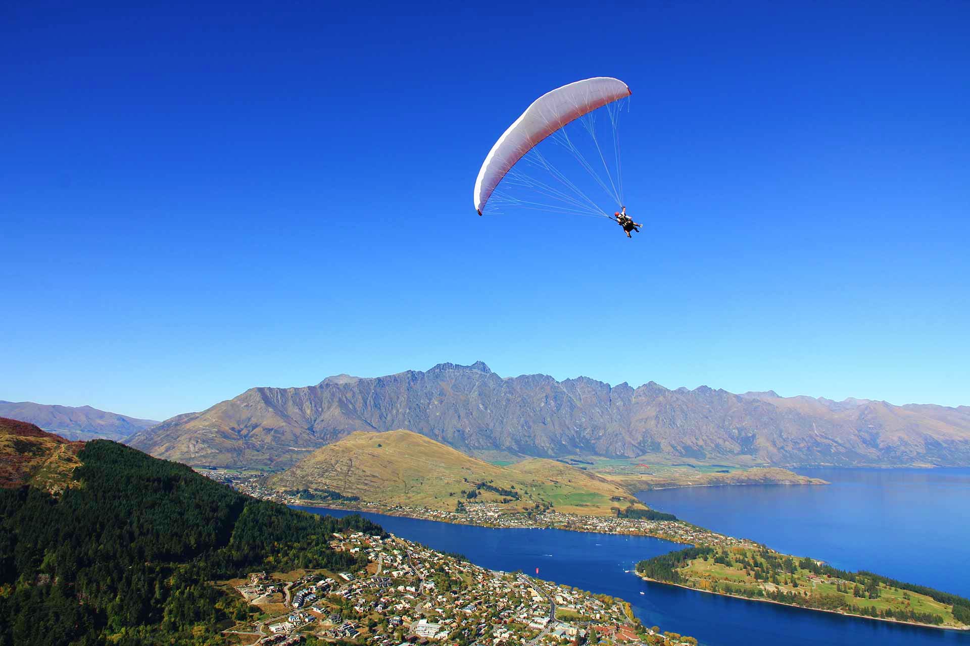 Paraglider soaring above Lake Wakatipu with Queenstown and The Remarkables mountain range in New Zealand